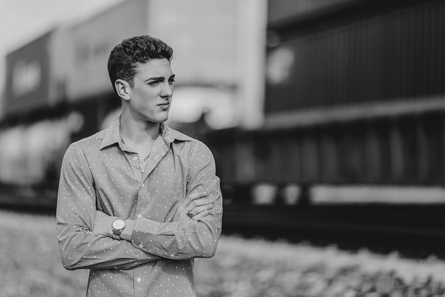Young man in striped shirt with arms crossed standing near railroad tracks in black and white photo.