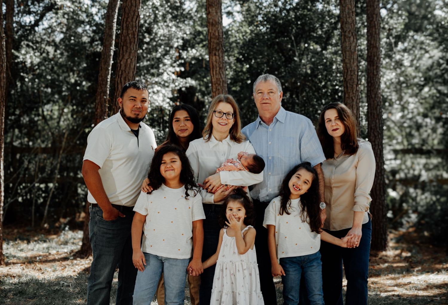 Large multigenerational family portrait outdoors among tall pine trees, including grandparents, parents, and young children.