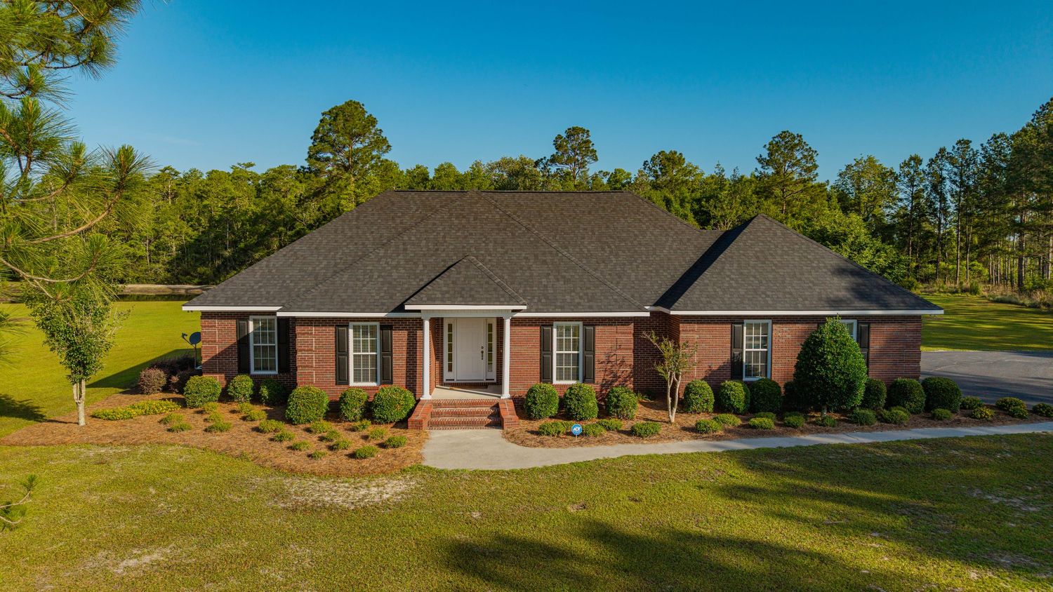 Brick ranch-style home with dark roof, manicured lawn, and lush green trees under a clear blue sky.