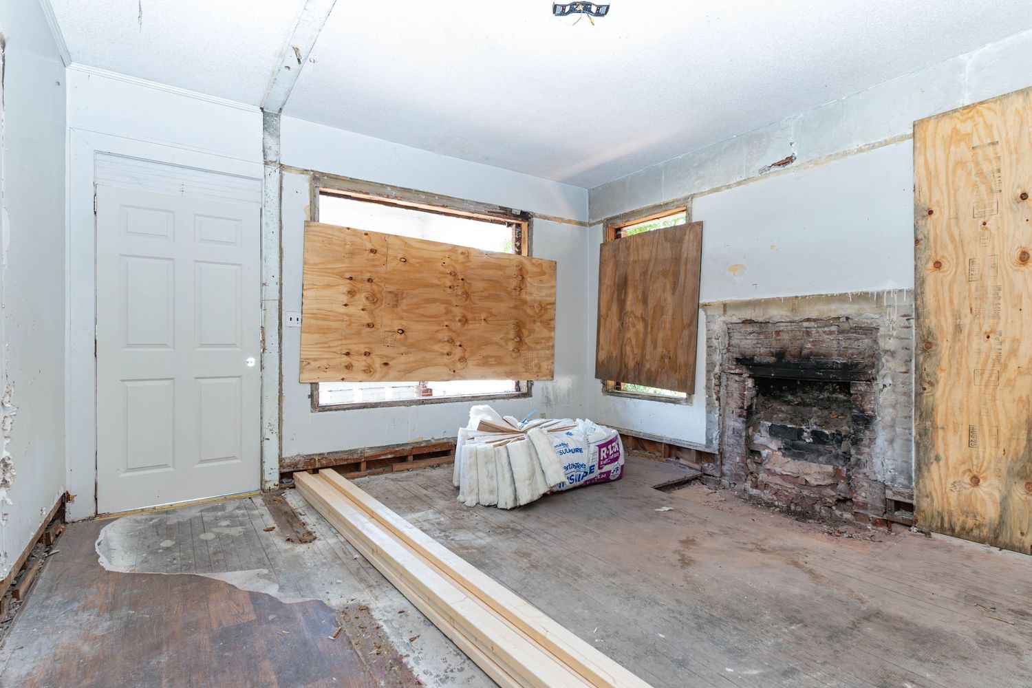 Vacant room under renovation with boarded windows, exposed fireplace, and construction materials on bare concrete floor.