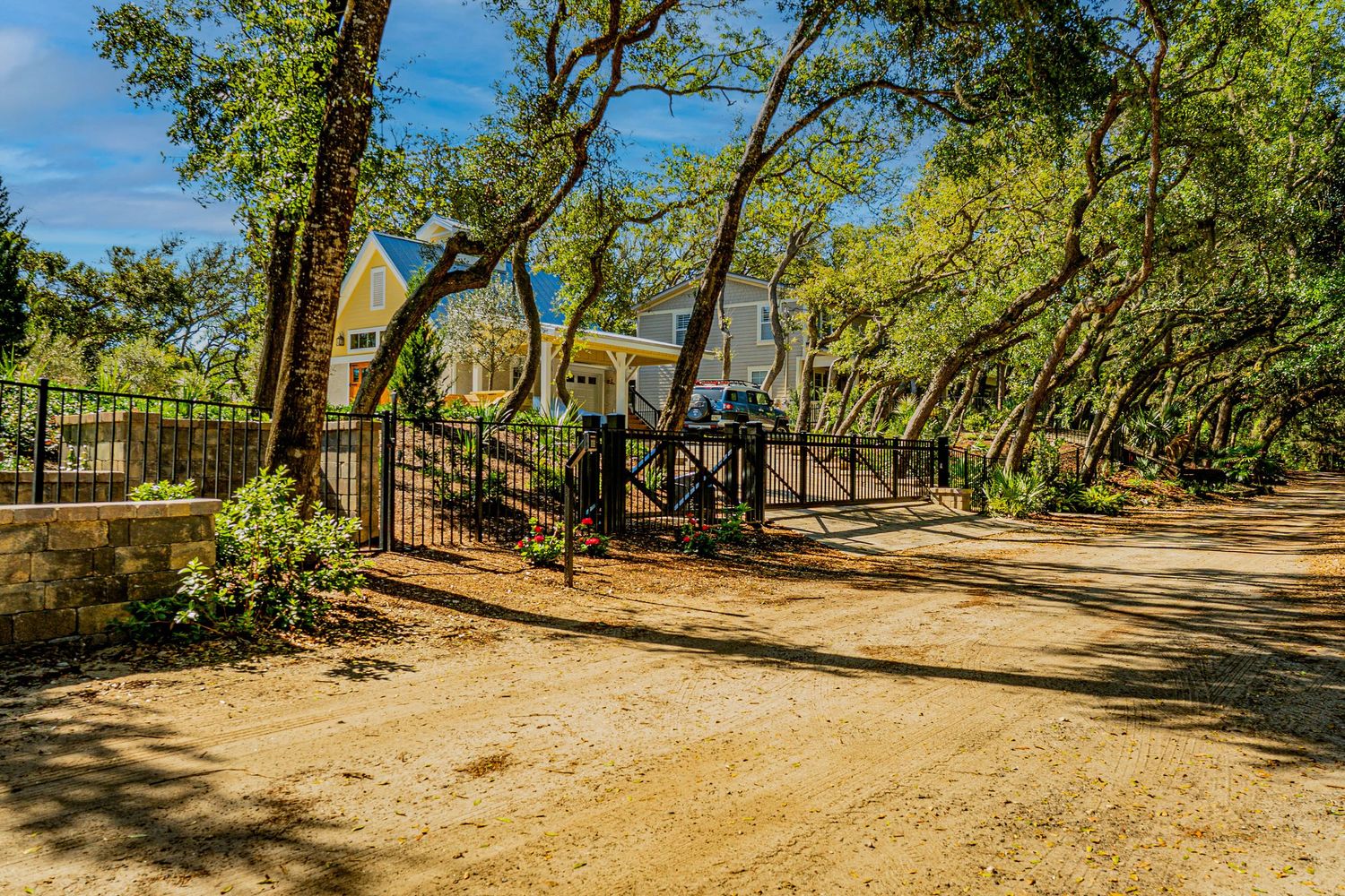 Gated gravel driveway entrance surrounded by mature oak trees leading to a charming yellow house in a wooded setting.