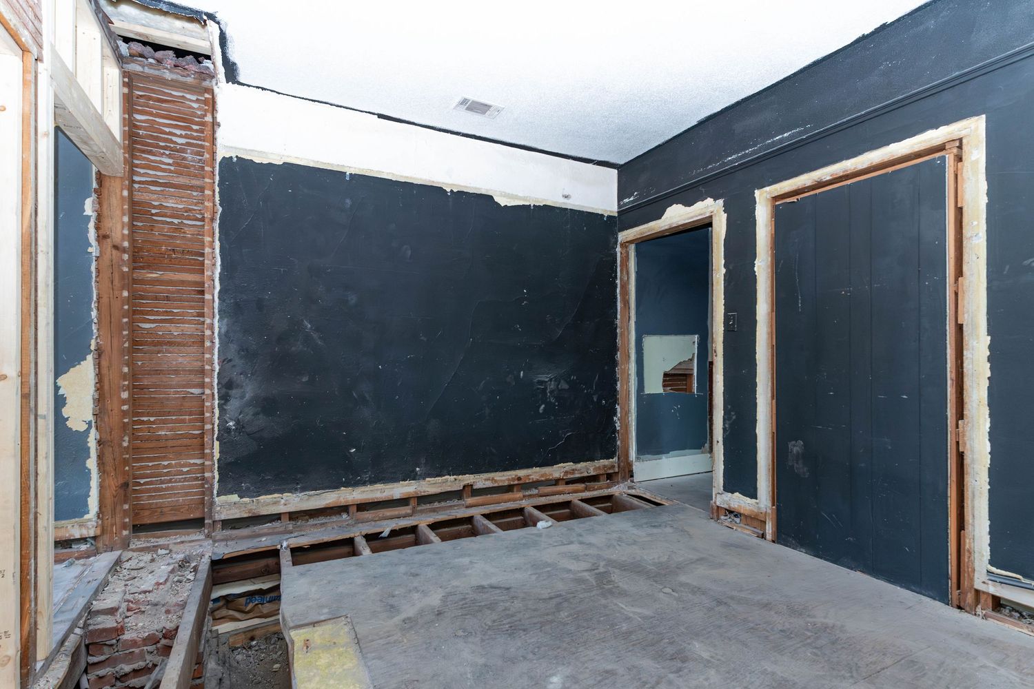 Interior of a room under renovation with exposed wall framing, black walls, and bare concrete floors.