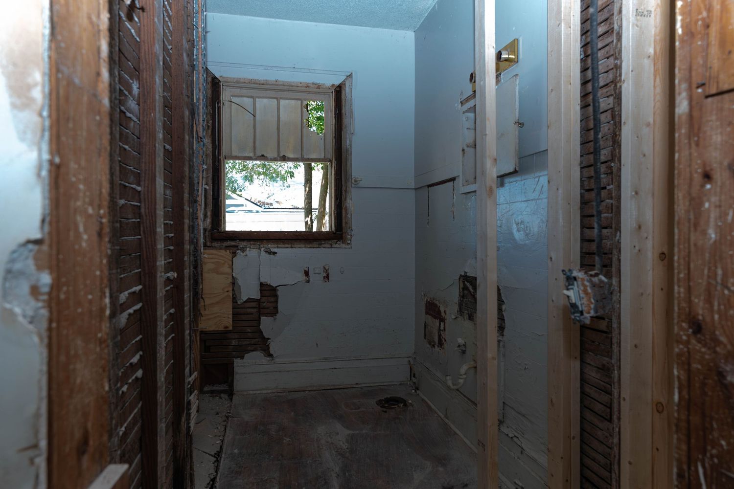 Interior view of a home under renovation showing exposed wall studs, damaged drywall, and a small window at the end of a hallway.