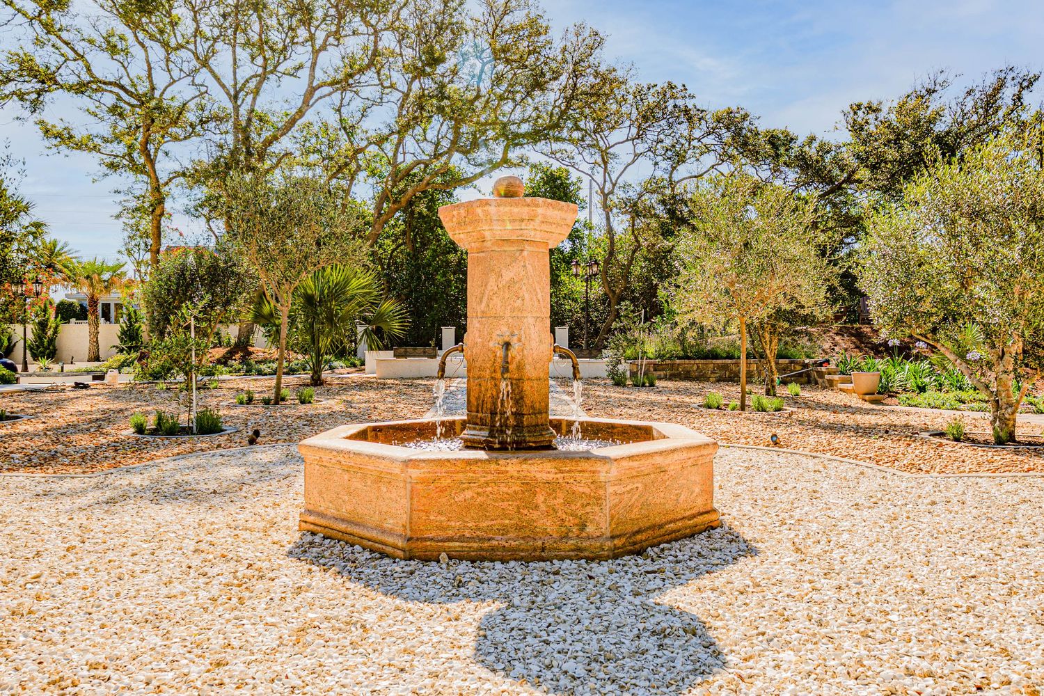 Stone fountain with cascading water surrounded by gravel pathways and lush trees in a serene garden setting.