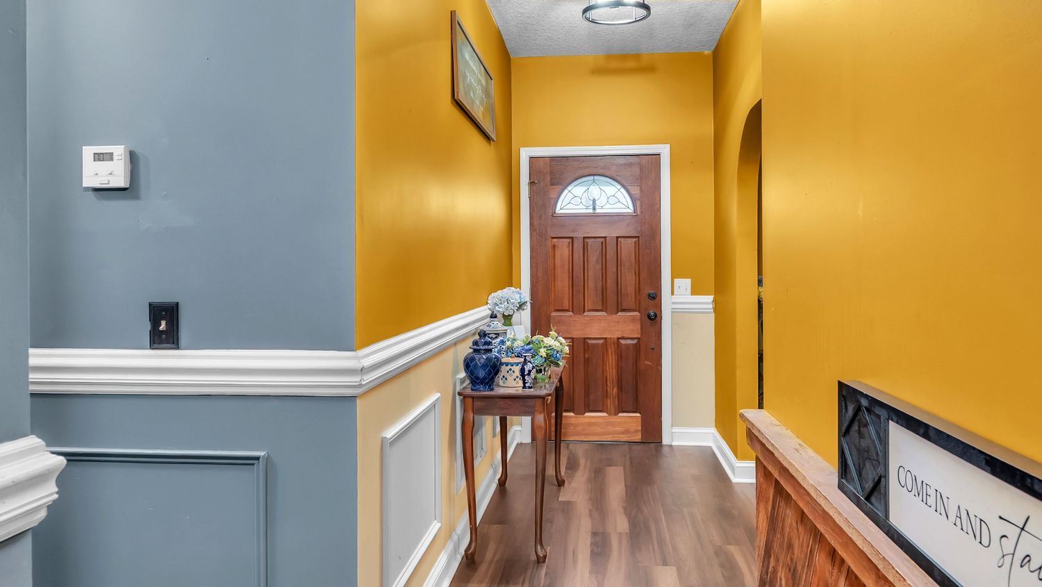 Bright entryway with yellow walls, wainscoting, hardwood floors, and a wooden front door with a sunburst window.