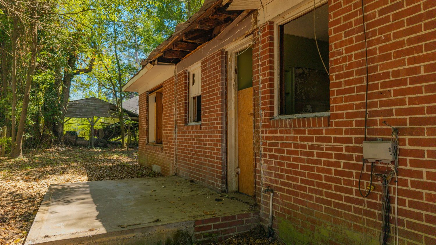 Abandoned brick building with missing windows and doors, surrounded by overgrown trees and a concrete pathway.