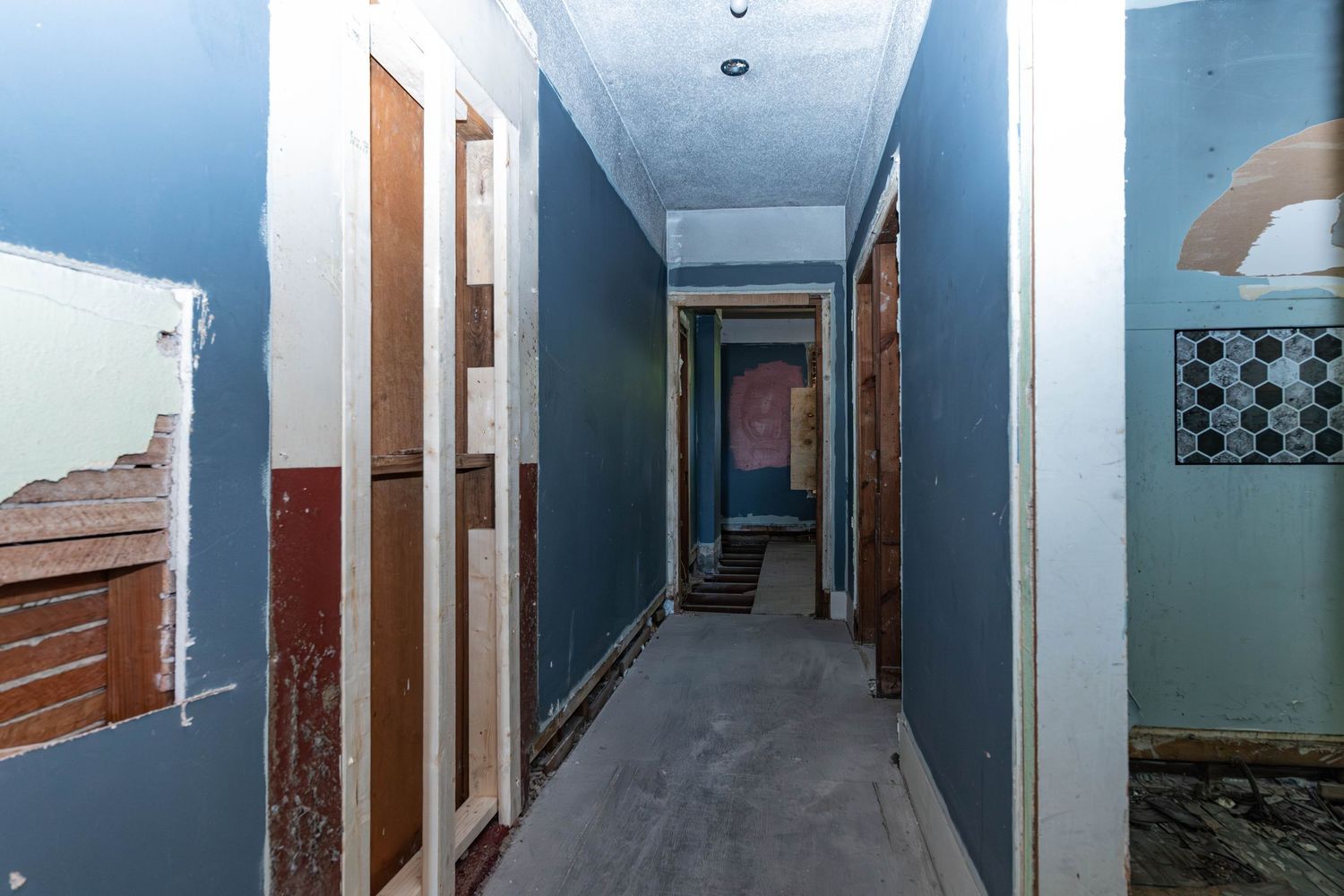Narrow hallway under renovation with blue walls, exposed door frames, and concrete floors in a gutted interior space.