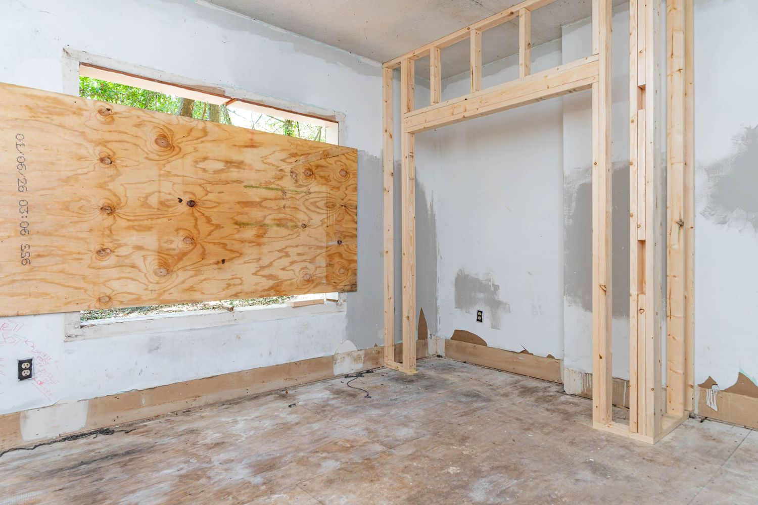 Empty room mid-renovation featuring plywood-covered window, new wood stud framing, and bare concrete floor.
