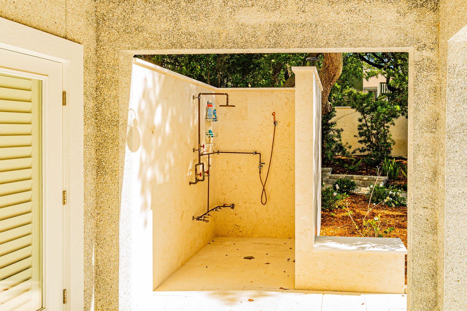 Outdoor shower enclosure with exposed pipes set within a stone archway surrounded by lush greenery.