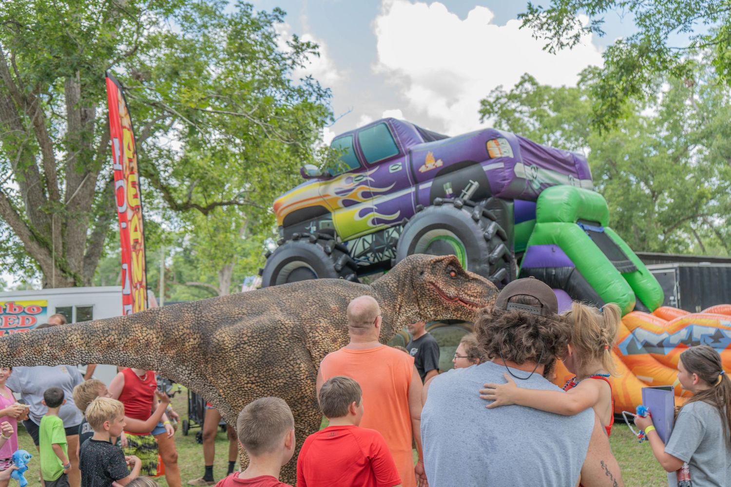 Kids and adults watch a large purple monster truck display at an outdoor fair with inflatable attractions nearby.