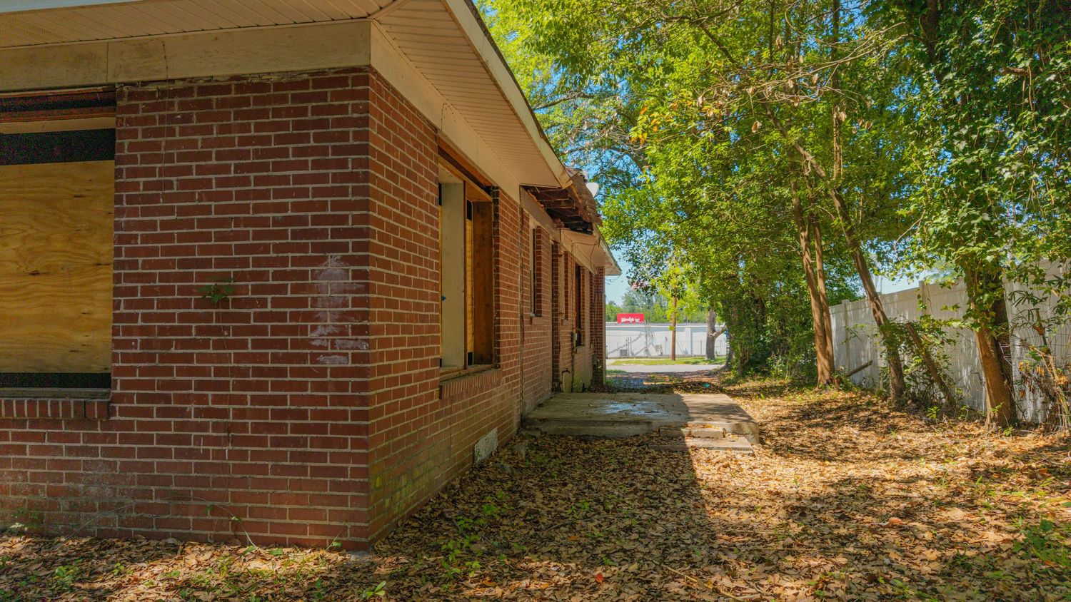 Abandoned brick building with boarded windows surrounded by overgrown trees and fallen leaves on a sunny day.