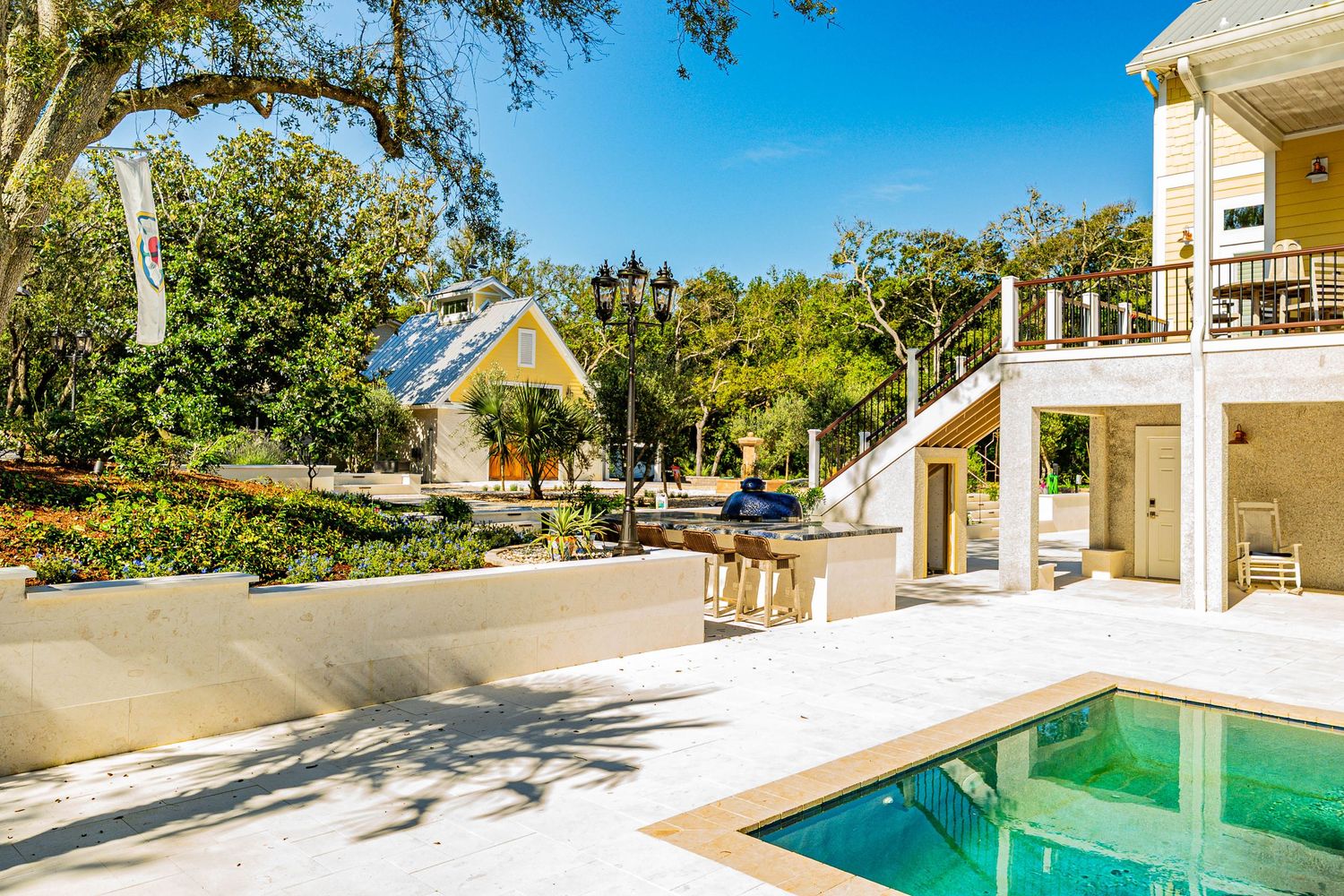 Luxury home with a sparkling pool, lush trees, yellow cottage, and elegant staircase on a sunny day.