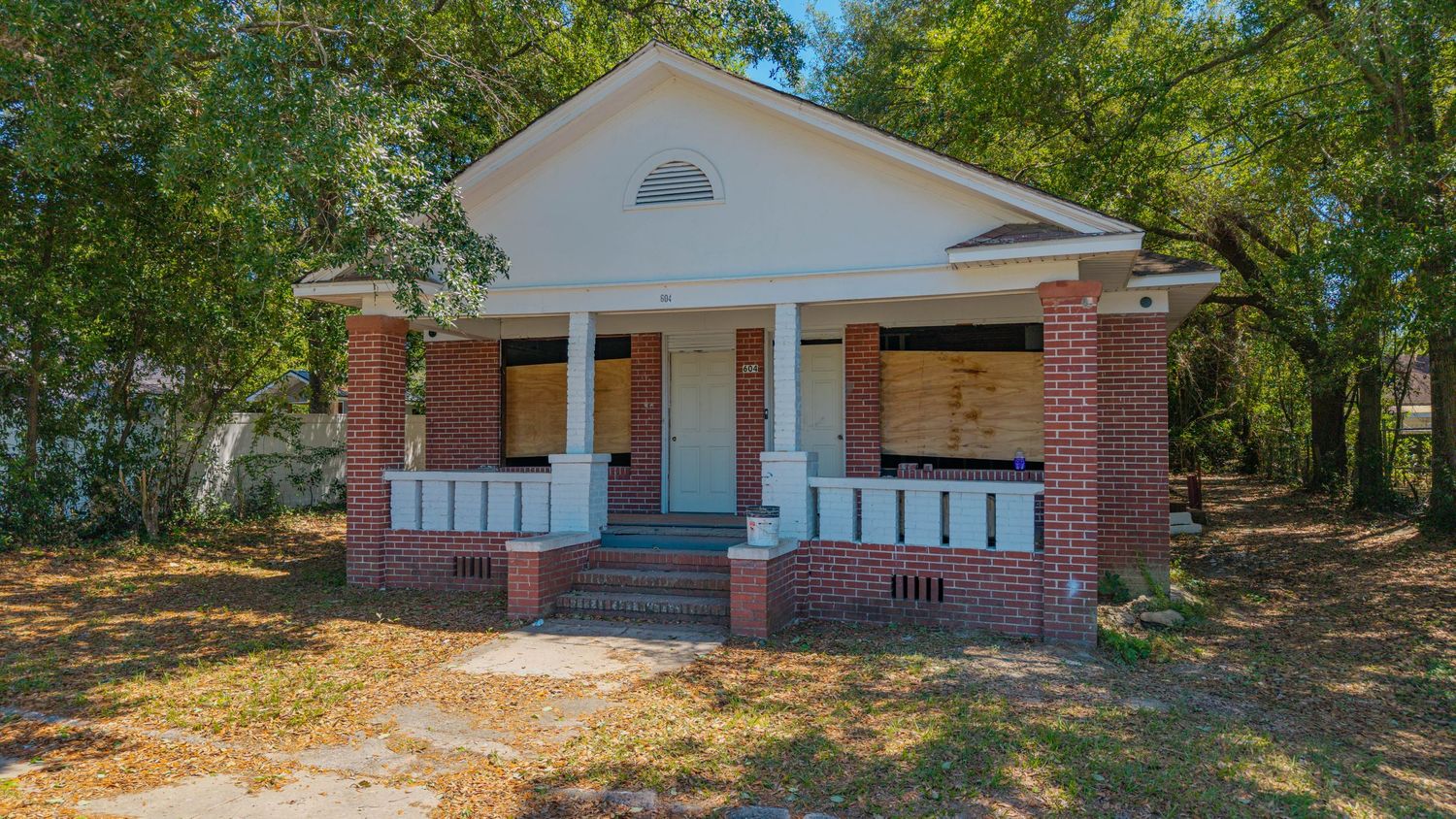 Abandoned brick bungalow with boarded windows, covered front porch, and overgrown yard surrounded by tall trees.