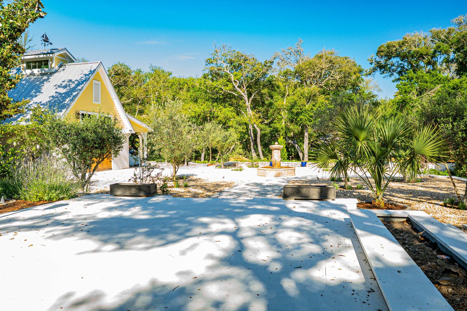 White sandy courtyard with palm trees and a charming yellow cottage surrounded by lush green trees on a sunny day.