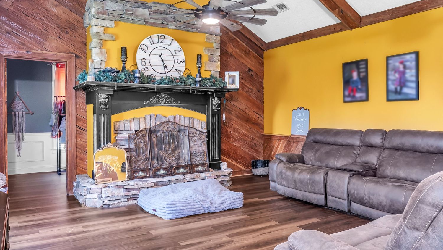 Cozy living room with stone fireplace, large clock, yellow walls, wood paneling, and reclining sofa on hardwood floors.