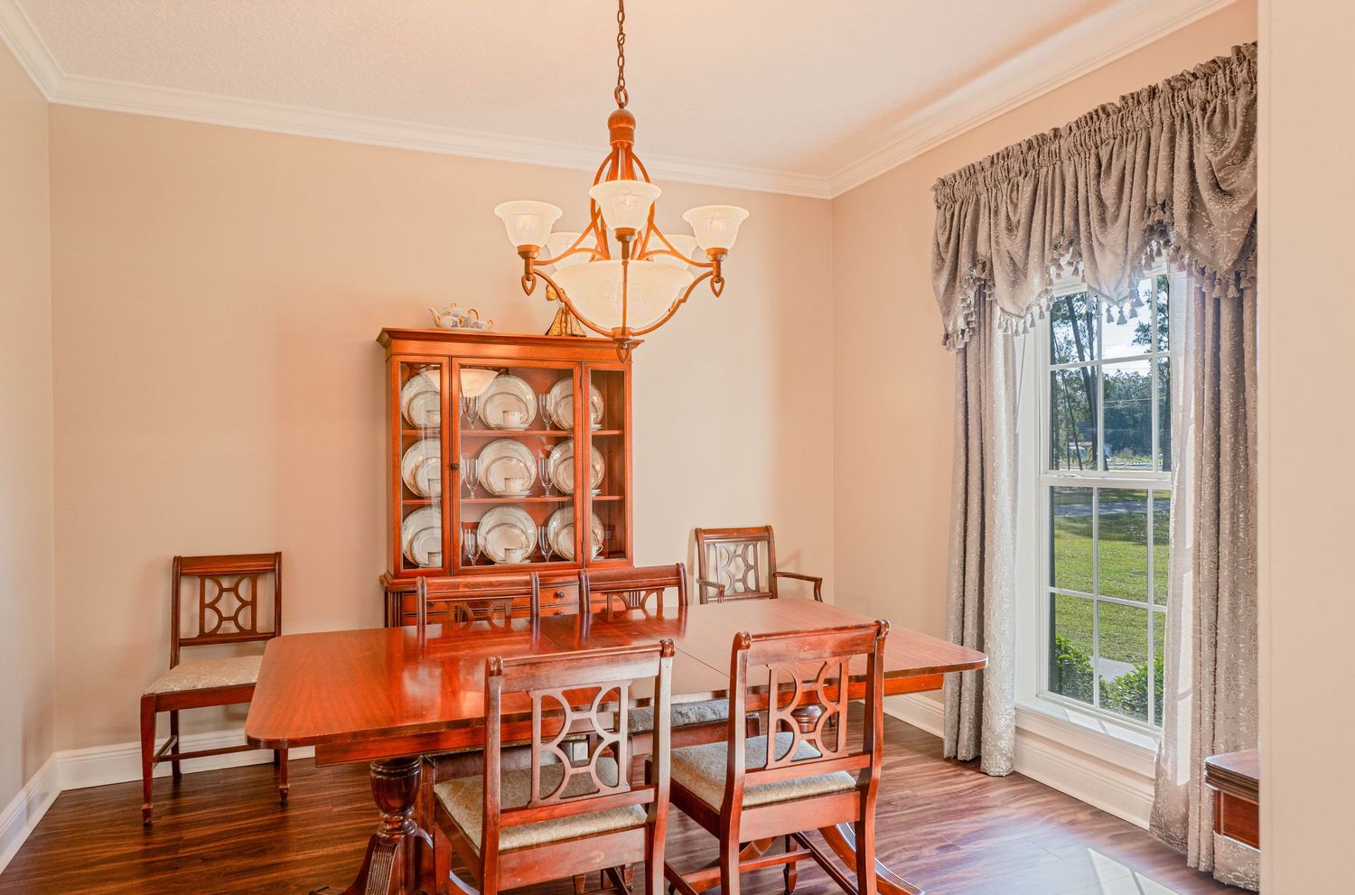 Elegant dining room with cherry wood table, chairs, china cabinet, hardwood floors, chandelier, and large window with drapes.
