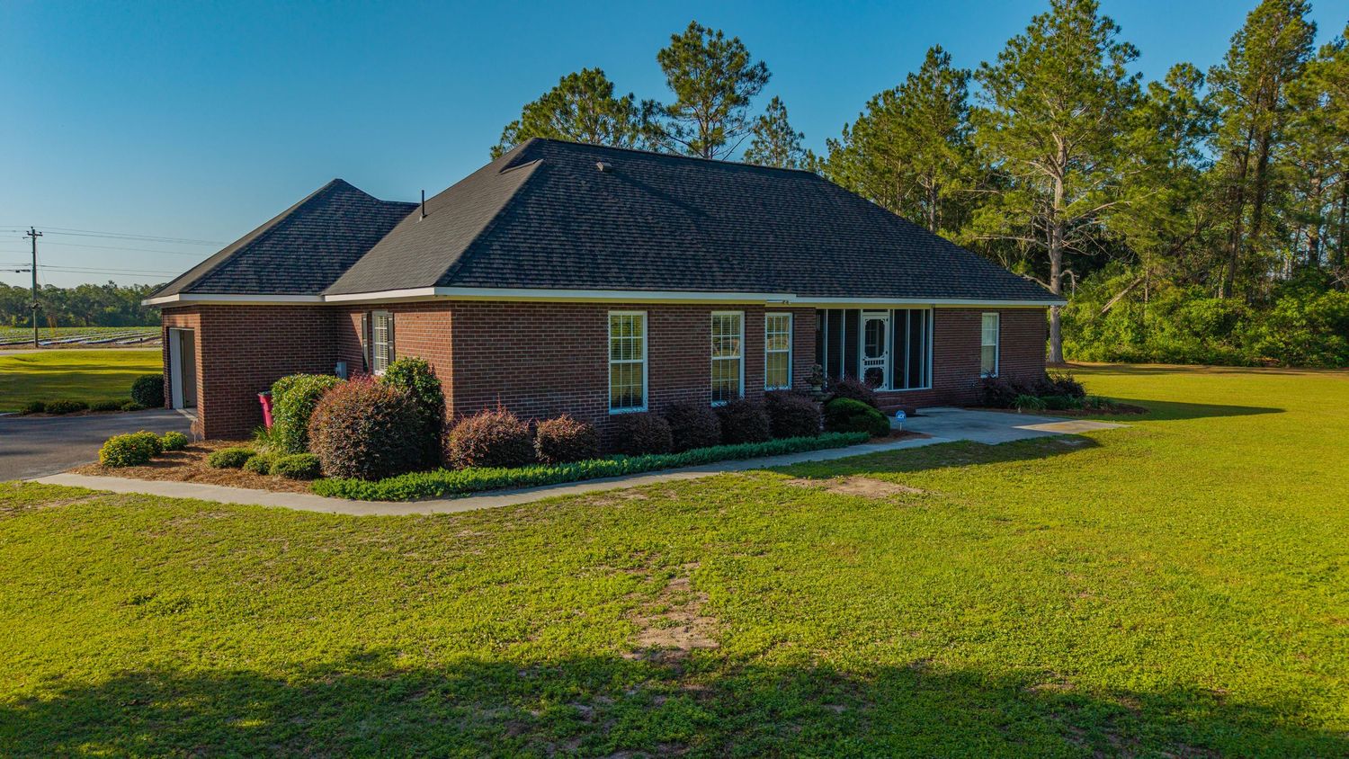 Brick ranch-style home with dark shingled roof, manicured green lawn, and mature pine trees under a clear blue sky.