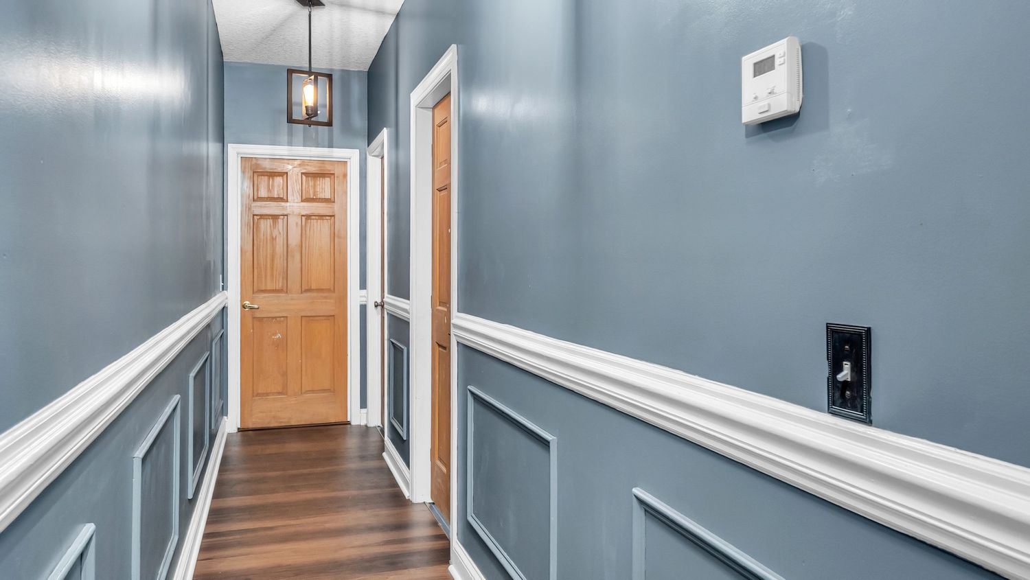 Blue hallway with white wainscoting, hardwood floors, and wooden doors with pendant lighting.