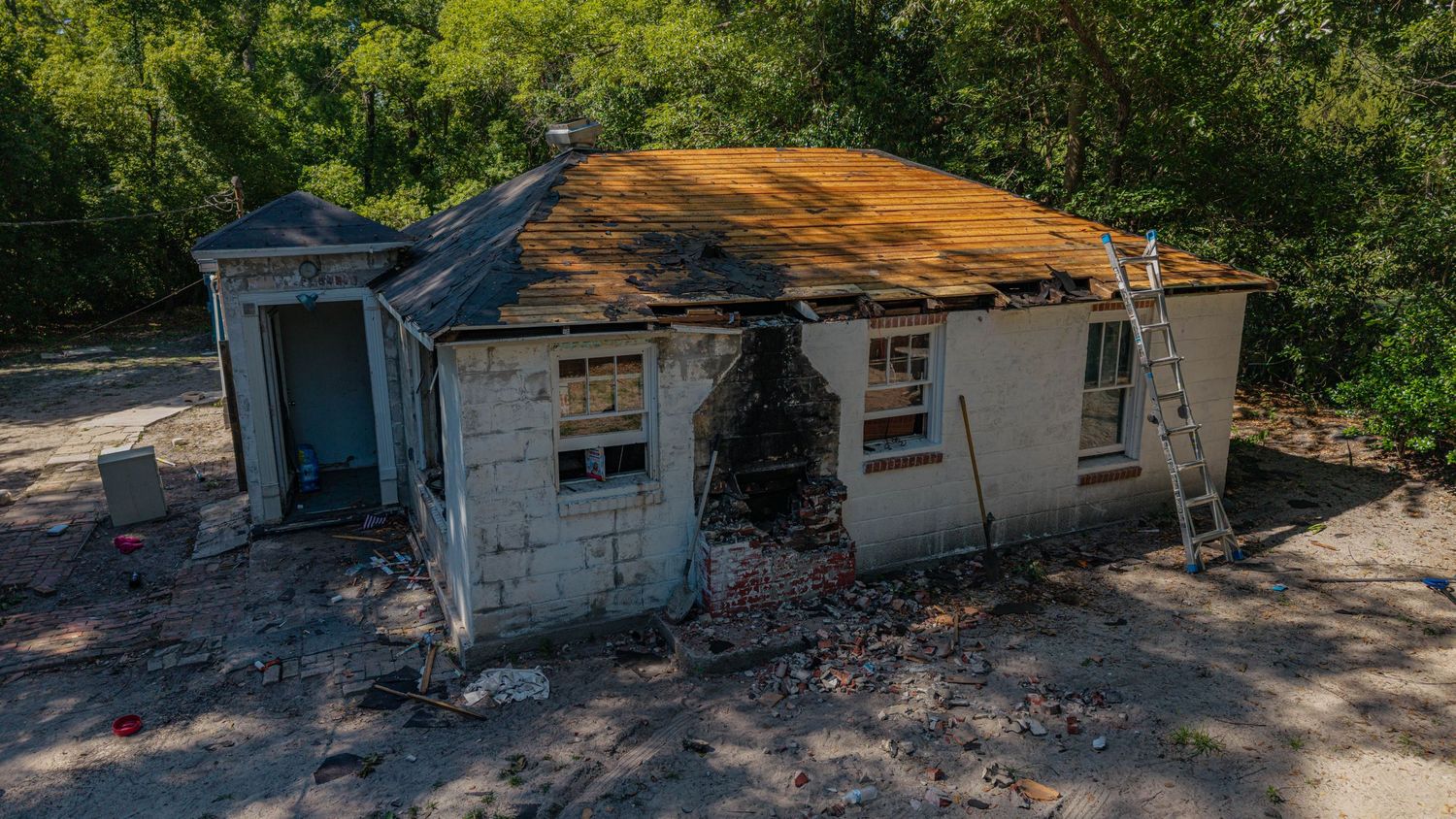 Fire-damaged abandoned house with charred walls, deteriorating roof, and debris scattered around the exterior.
