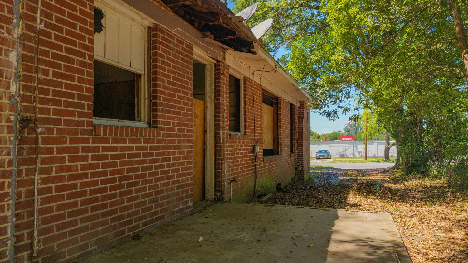Abandoned brick building with broken windows, overgrown trees, and a cracked pathway leading to a distant waterway.