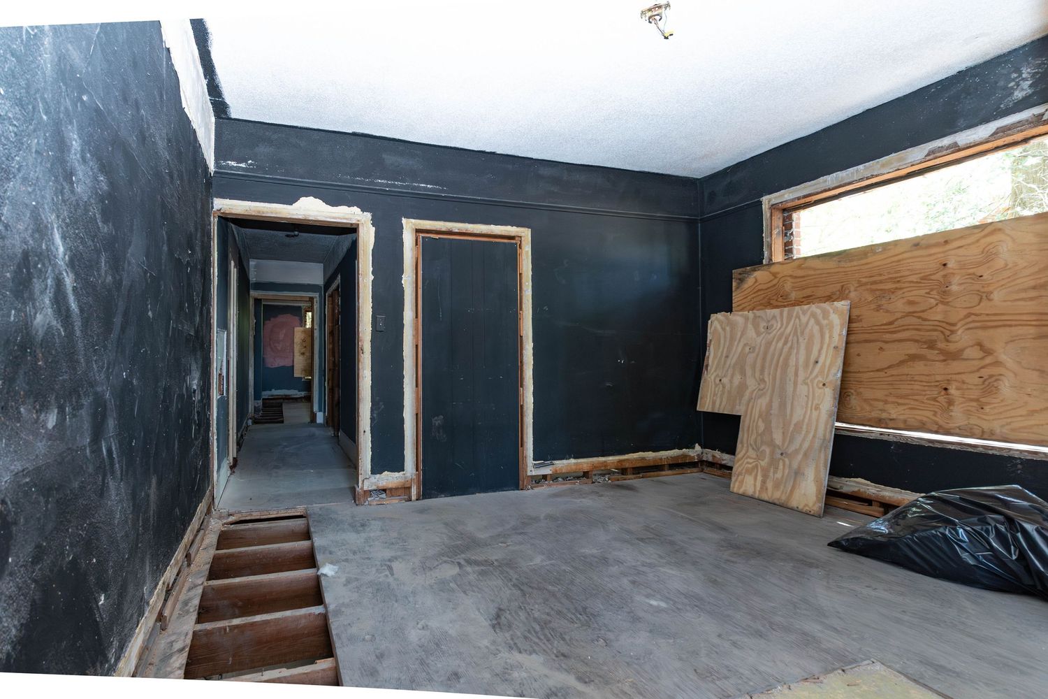 Abandoned room with dark walls, plywood-covered windows, concrete floor, and a hallway visible in the background.