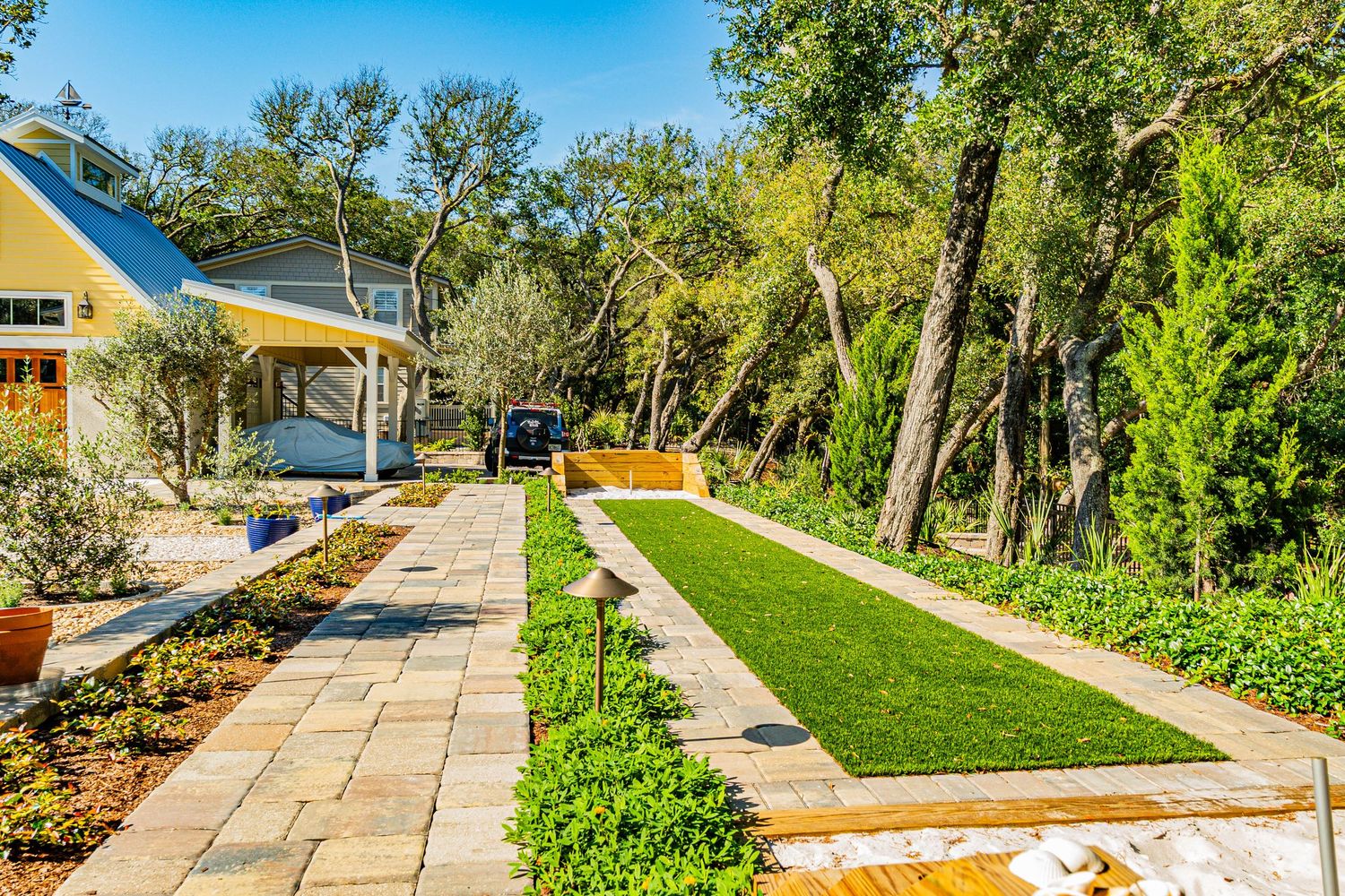 Landscaped backyard with bocce ball court, stone pathway, lush greenery, and mature trees surrounding a yellow home.