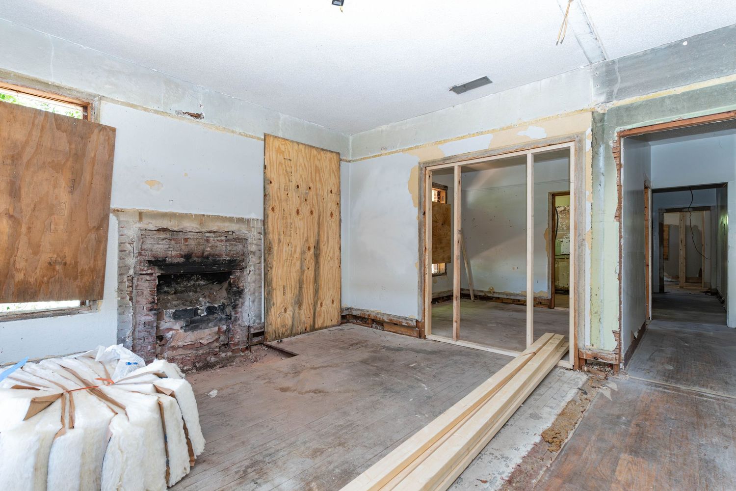 Gutted room with brick fireplace, boarded windows, exposed walls, and lumber on floor during home renovation.