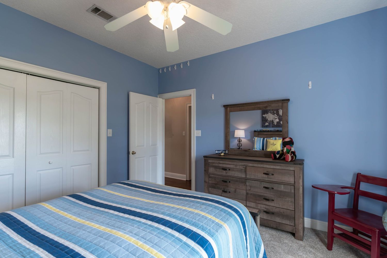 Cozy bedroom with blue walls, striped bedding, wooden dresser with mirror, ceiling fan, and white closet doors.