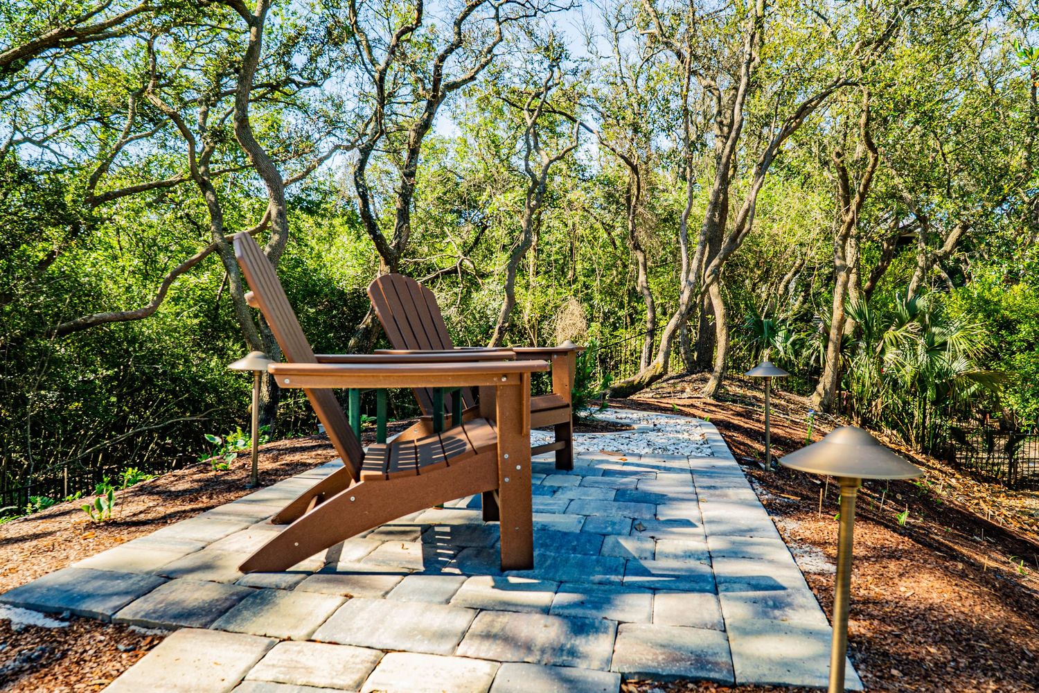 Wooden Adirondack chair on a stone patio path surrounded by lush green trees and landscaped garden.