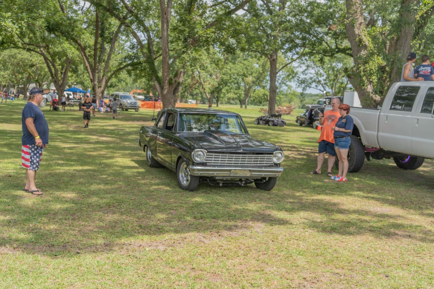 Vintage black car on display at an outdoor car show under large oak trees with spectators nearby.
