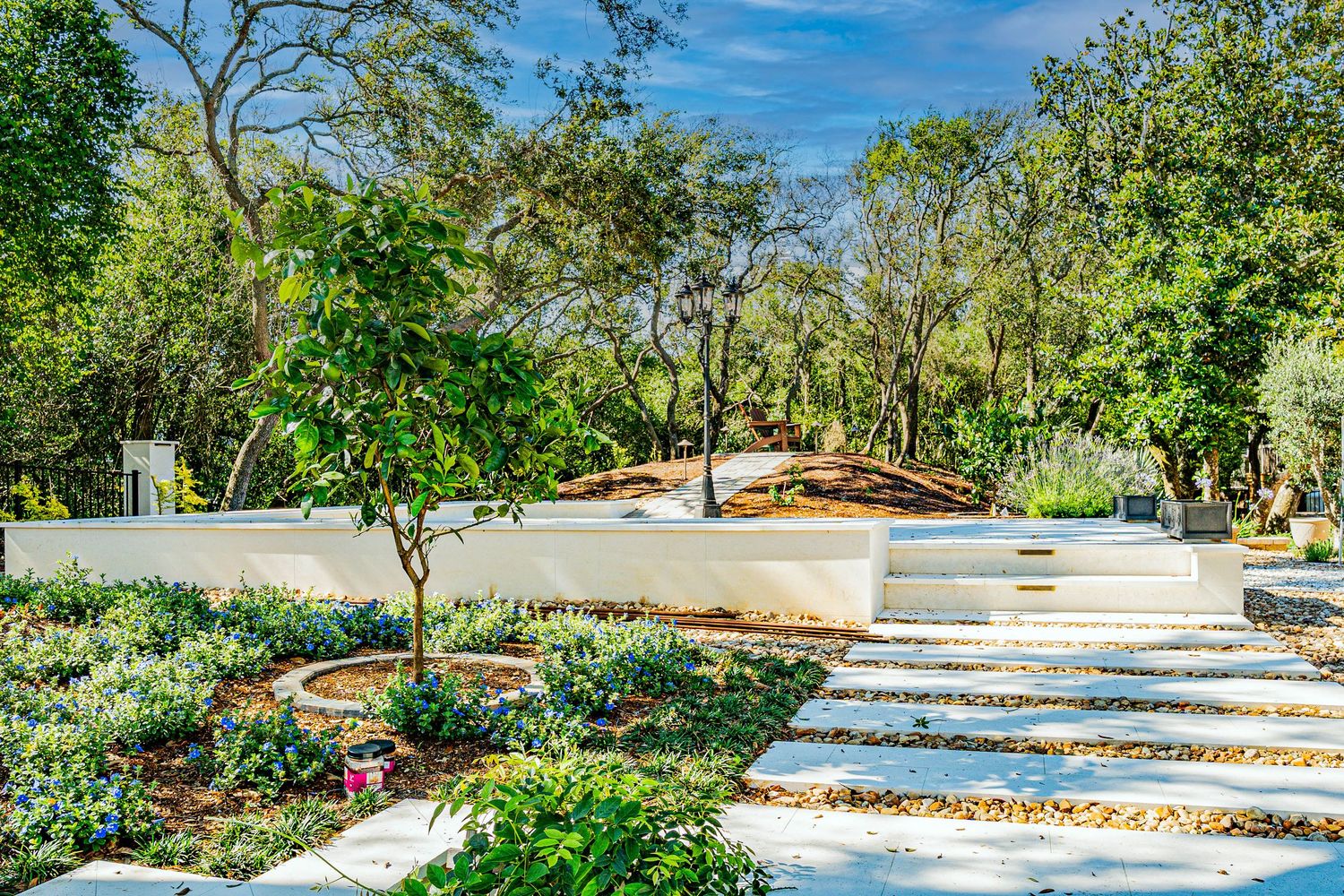 Lush garden landscape with white stone steps, flowering plants, and a curved wooden structure surrounded by tall trees.