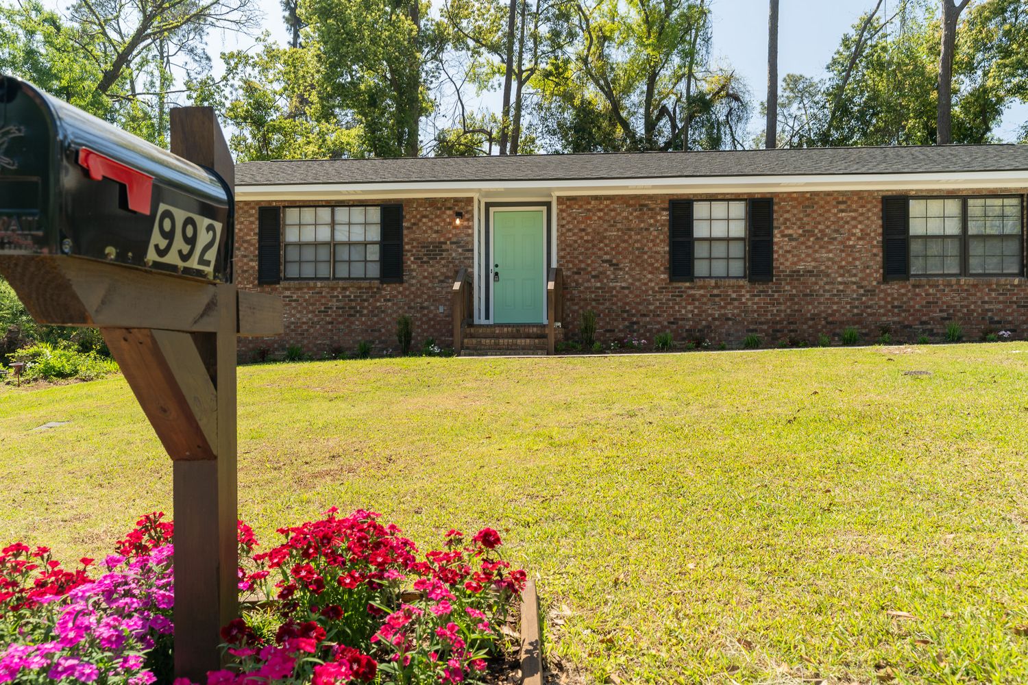 Brick ranch home at 992 with teal front door, green lawn, pink flowers, and mailbox in foreground.