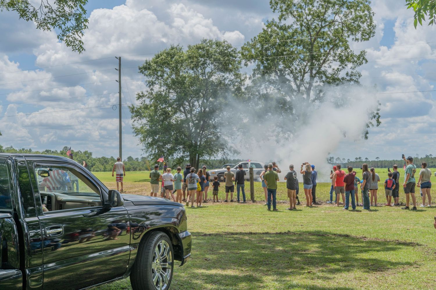 Crowd gathers outdoors near a black truck watching a smoke demonstration in a green field under a cloudy sky.