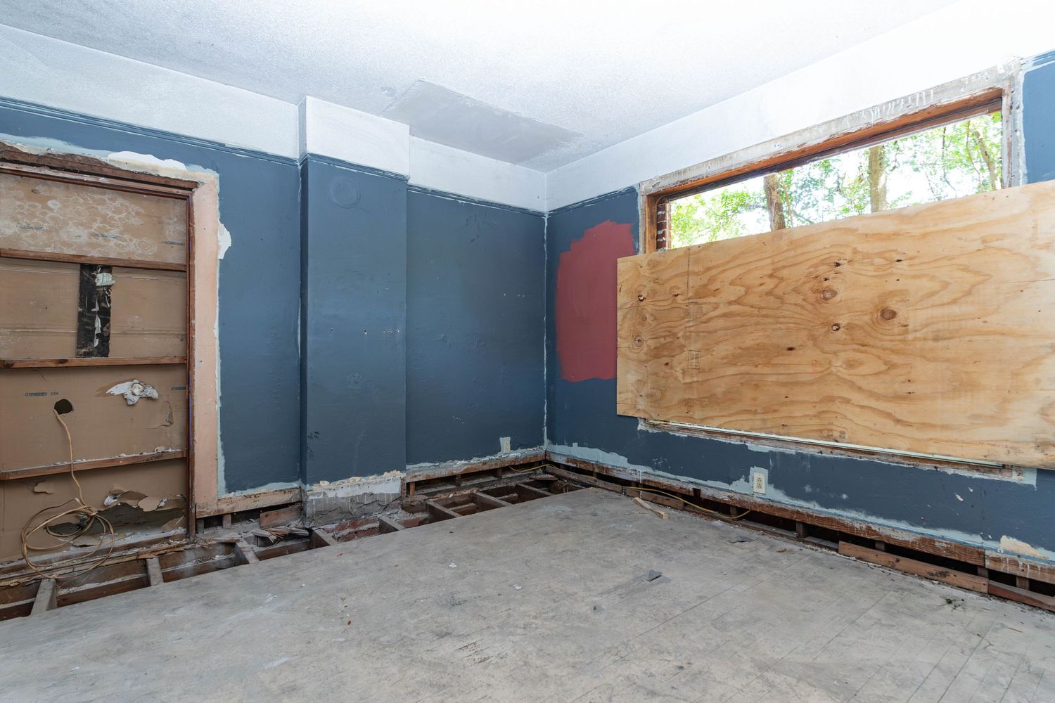 Gutted room with blue walls, boarded window, exposed concrete floor, and damaged drywall in a home under renovation.