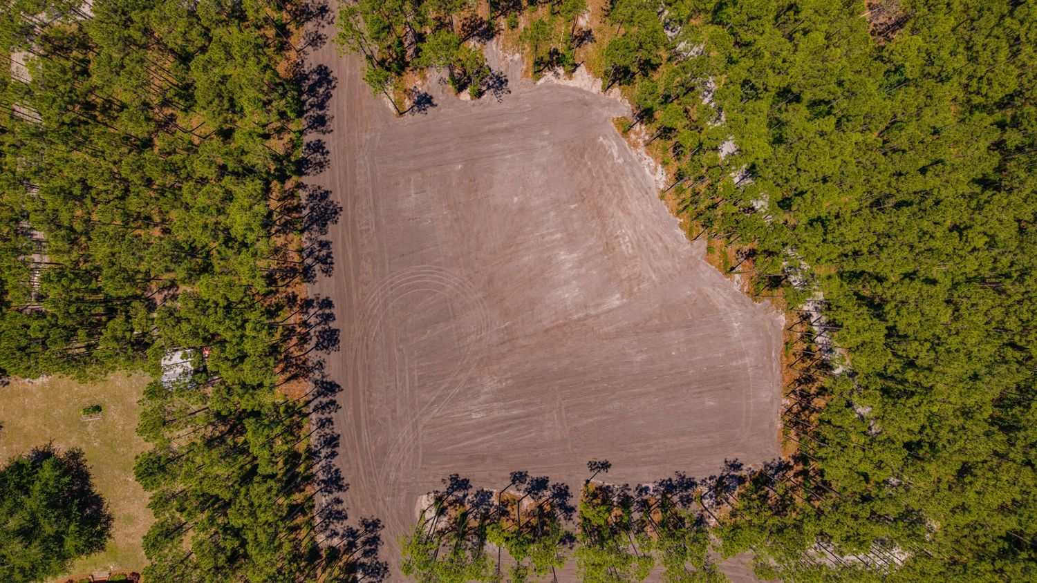 Aerial view of a large cleared dirt lot surrounded by dense green pine trees in a forested area.