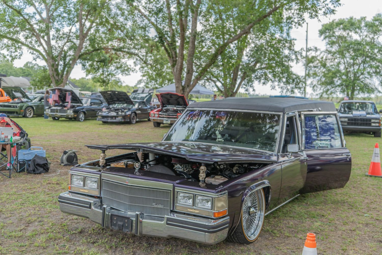 A lowrider classic black Cadillac hearse on display at an outdoor car show with trees in the background.
