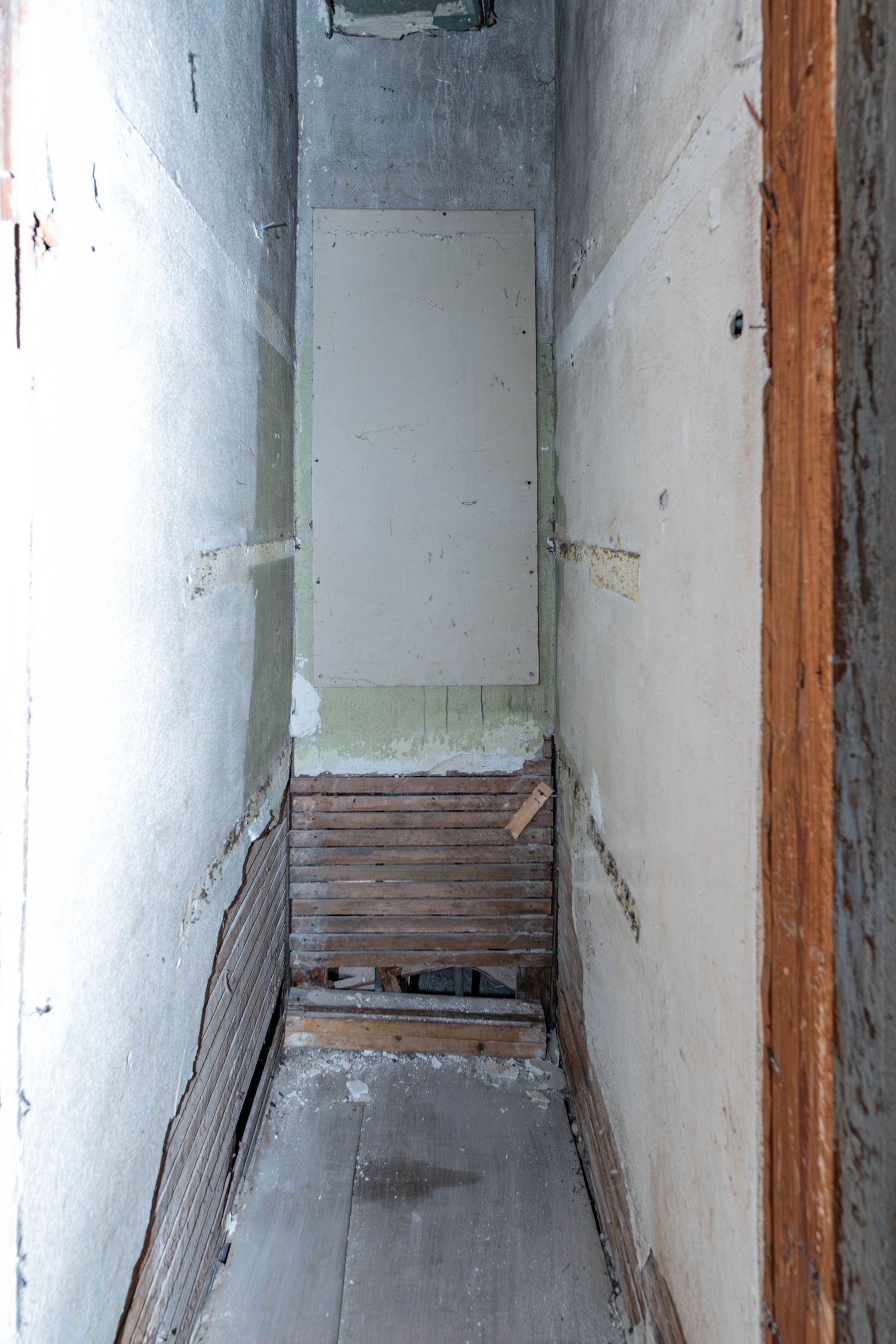 Narrow dilapidated hallway with peeling paint, exposed wood, and debris leading to a worn door under renovation.