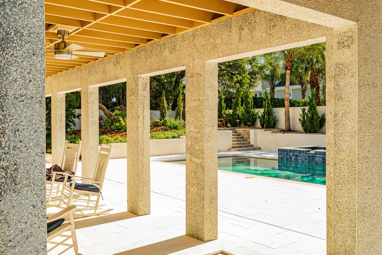 Covered patio with concrete columns overlooking a sparkling pool surrounded by lush greenery and lounge chairs.