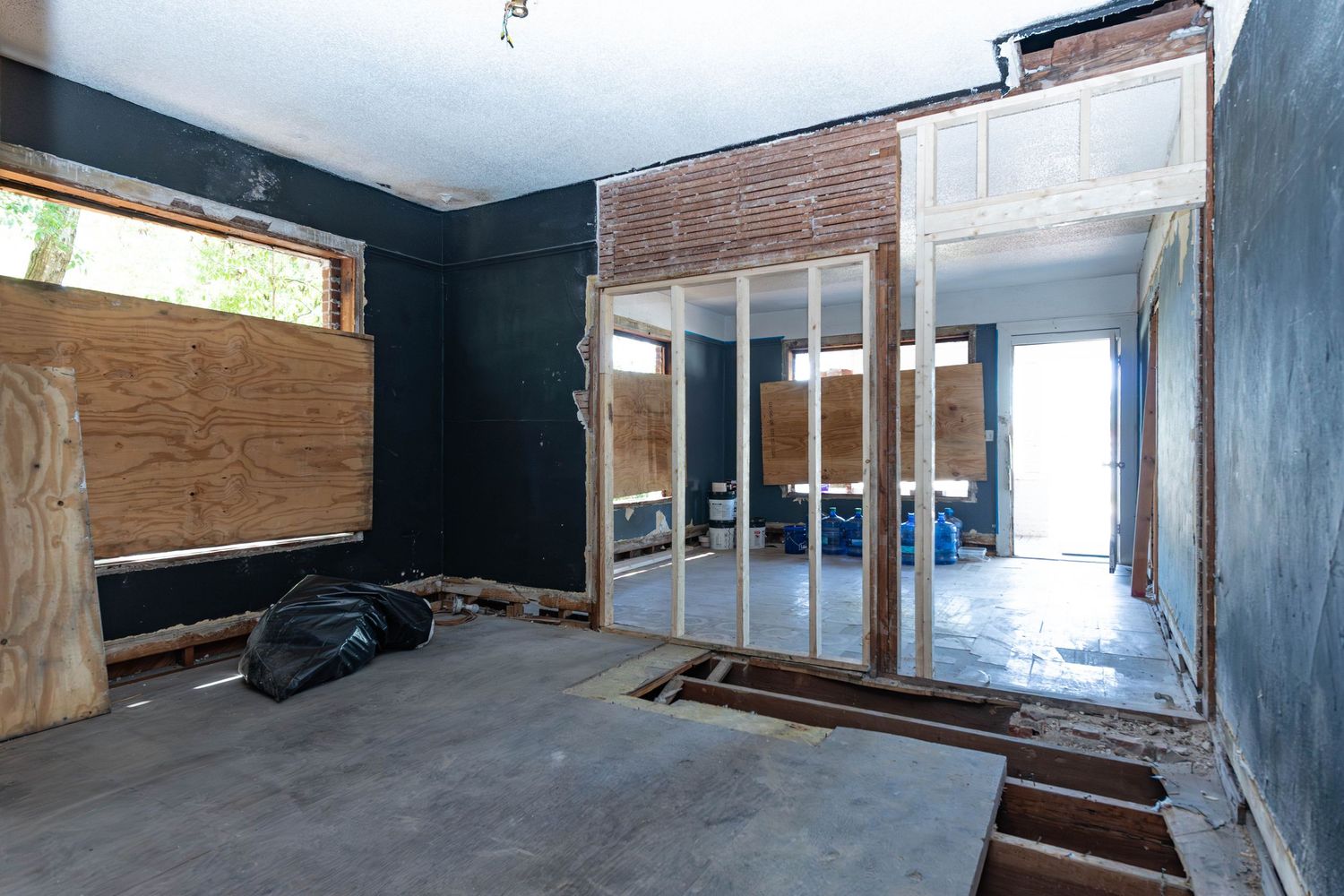 Gutted room interior showing plywood-covered walls, exposed brick, sliding glass doors, and damaged flooring during home renovation.