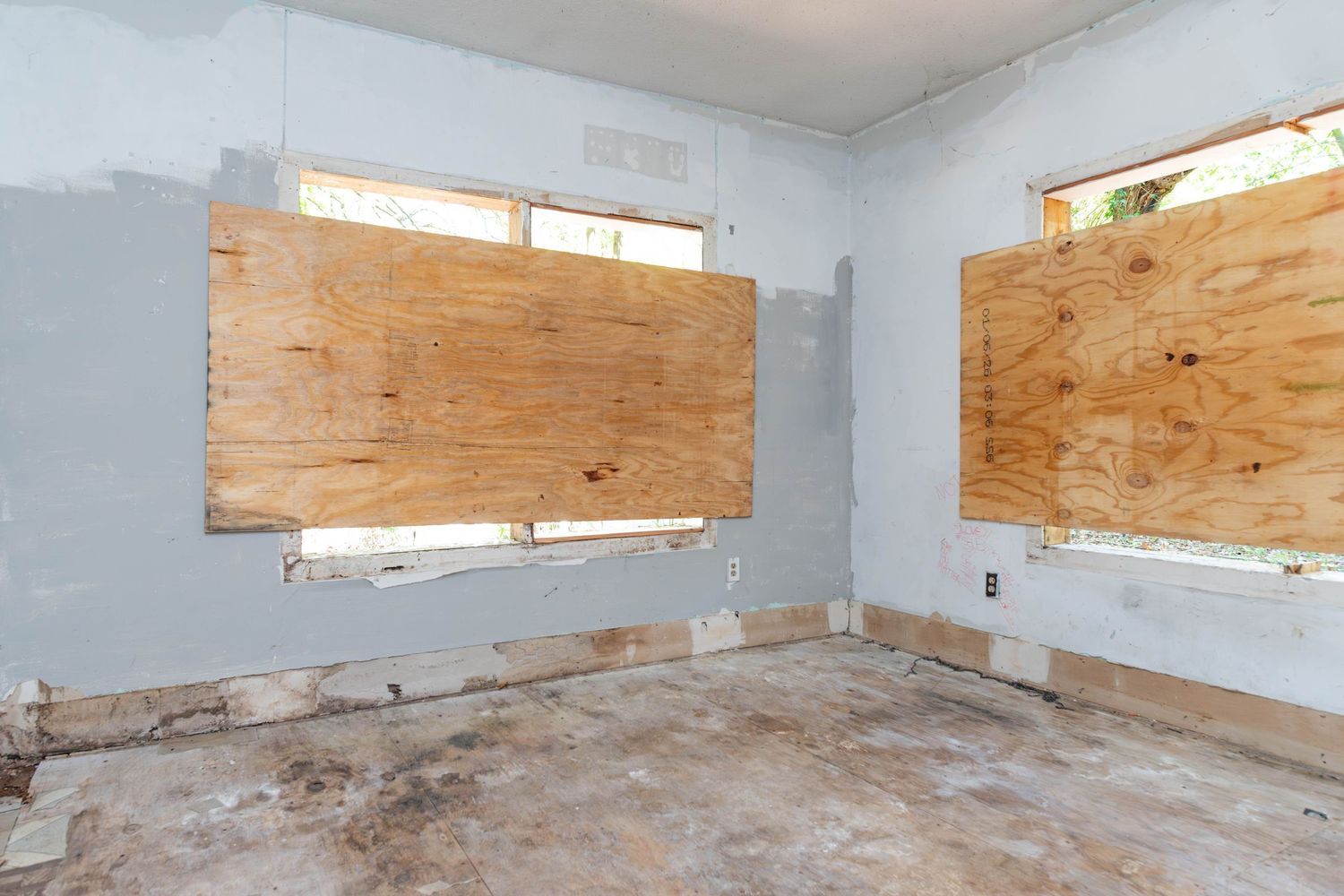 Abandoned room with boarded-up windows, stripped walls, and debris-covered floor undergoing renovation.