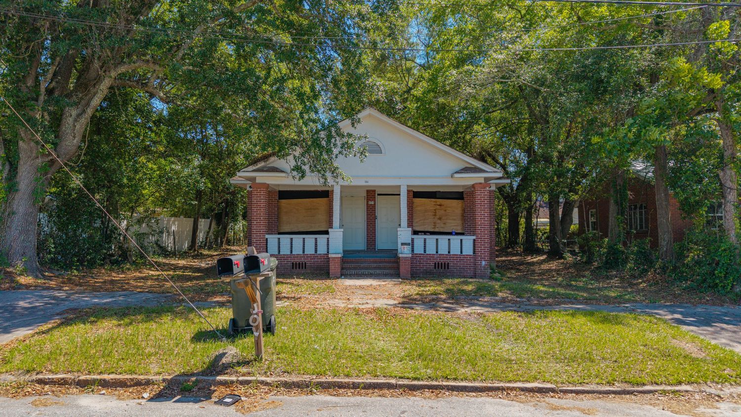 Abandoned brick bungalow with boarded windows, covered front porch, and overgrown lawn surrounded by large trees.