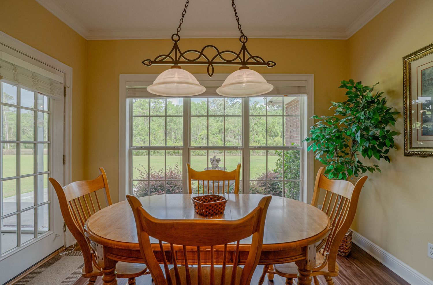 Bright dining room with oak table, Windsor chairs, chandelier, and large windows overlooking a lush green yard.