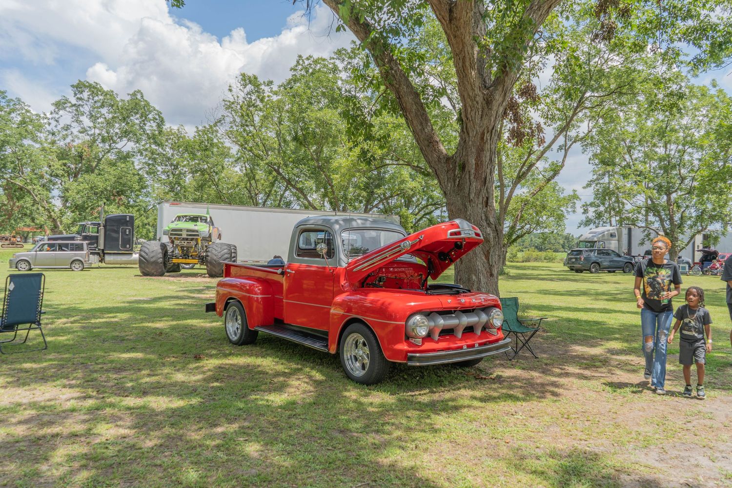 Vintage red Ford pickup truck with hood open displayed at outdoor car show under large tree.