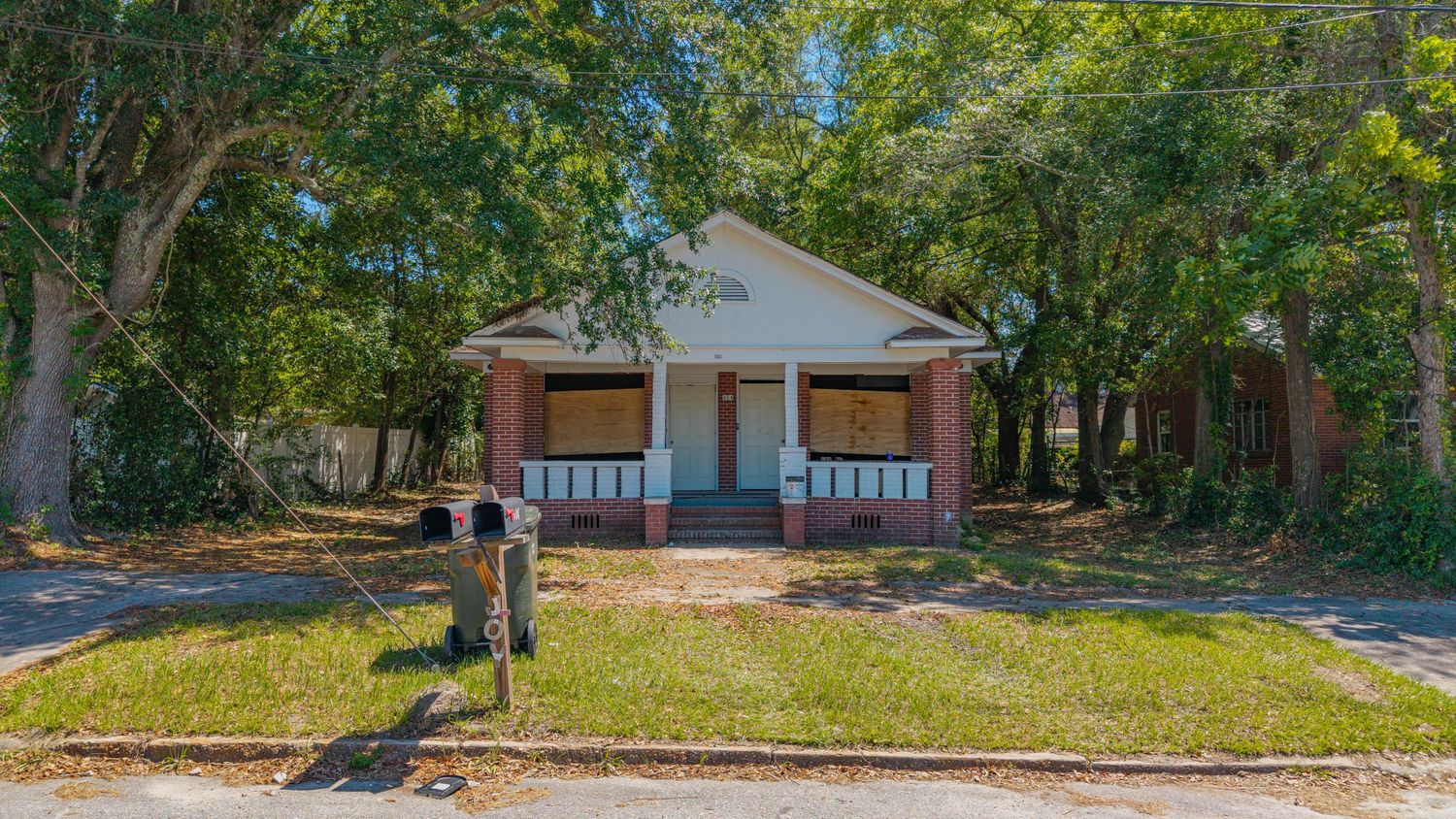 Abandoned brick bungalow with boarded windows, covered porch, and overgrown yard surrounded by large trees.