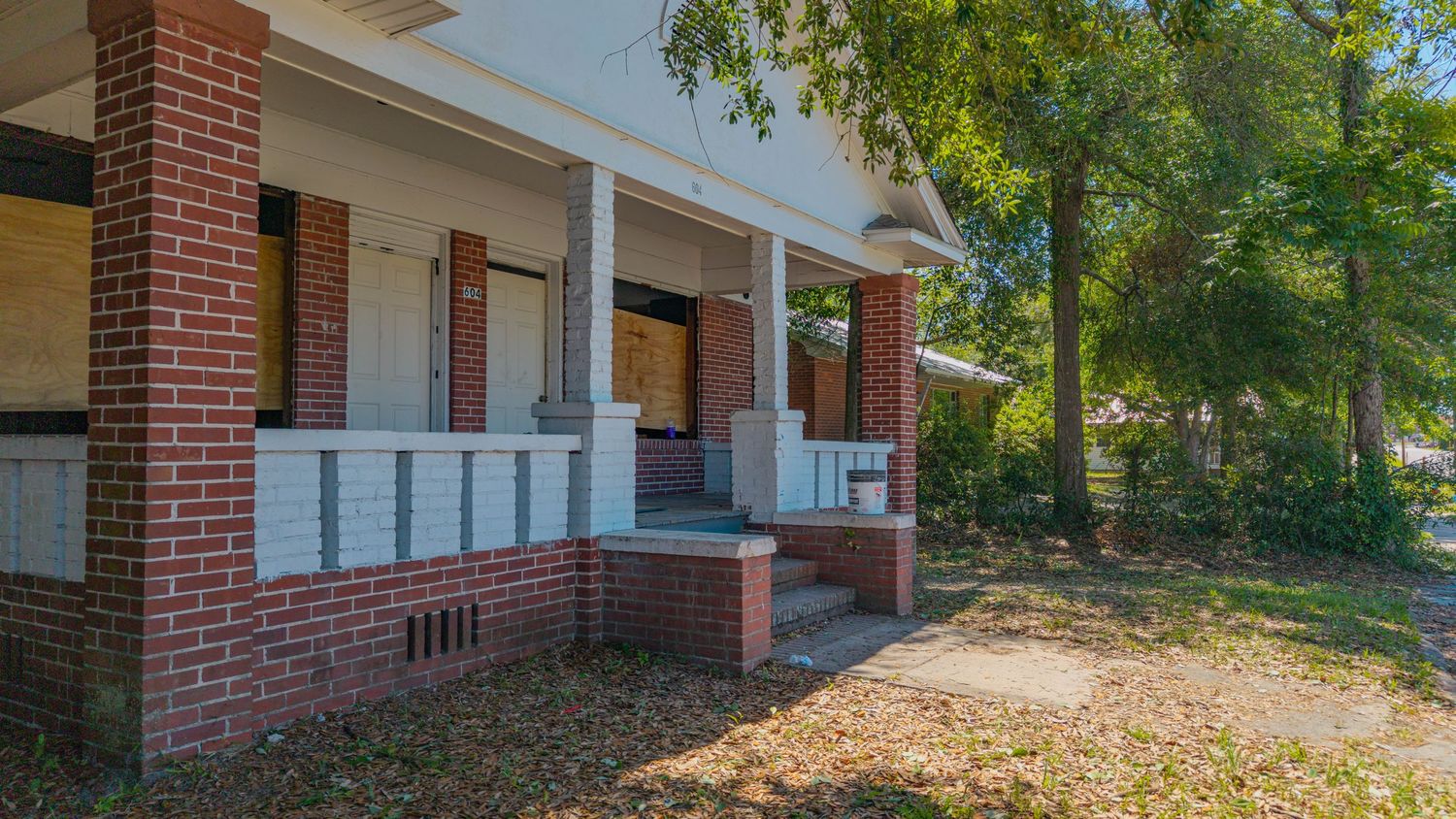 Abandoned brick house with boarded windows, covered front porch, and overgrown yard surrounded by trees.