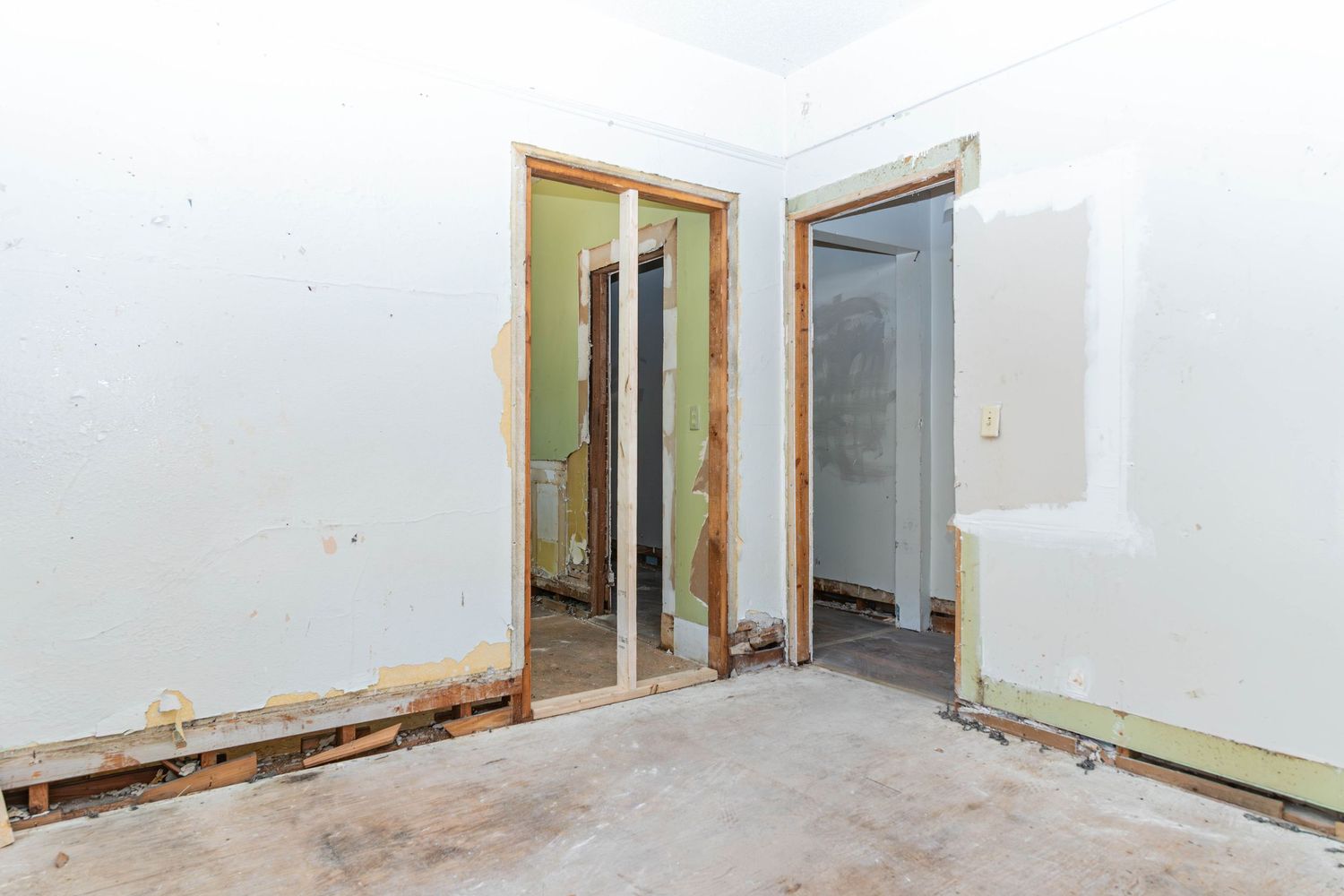 Unfinished room under renovation with bare concrete floors, exposed drywall, and two open doorways leading to adjacent spaces.