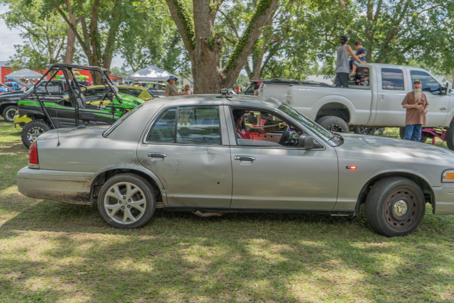 Silver Ford Crown Victoria sedan parked on grass at an outdoor car show with trucks and trees in background.