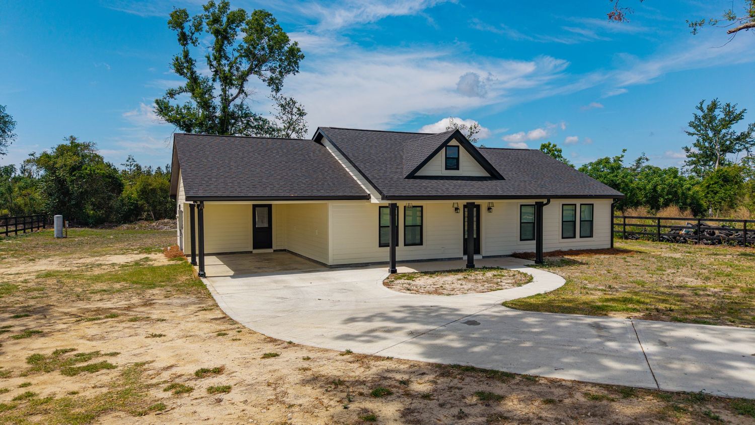 New single-story ranch home with dark roof, covered porch, and circular driveway surrounded by mature trees.