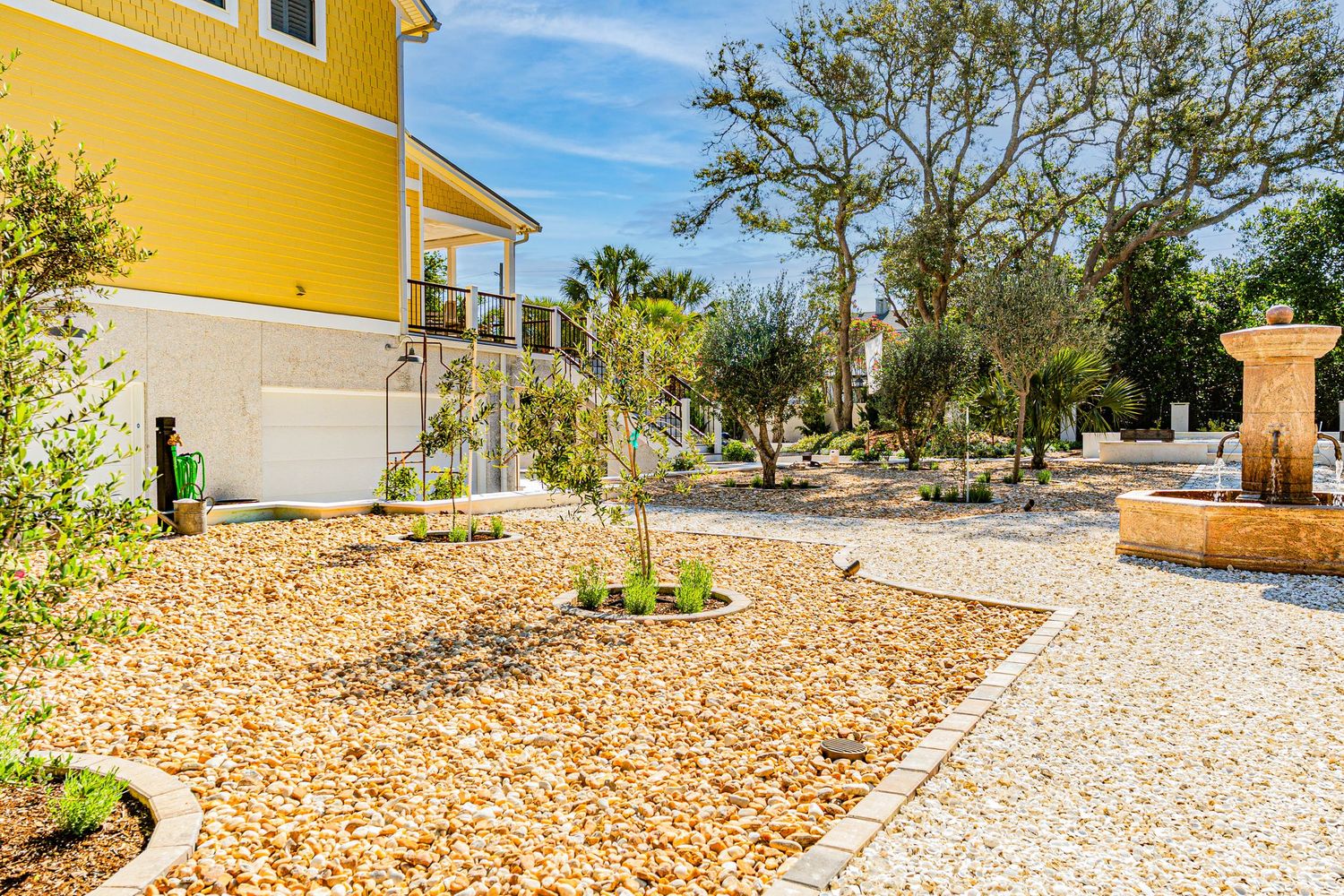 Yellow villa with gravel landscaping, olive trees, and stone fountain in a sunny Mediterranean garden.
