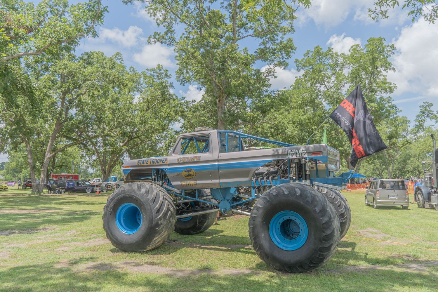 A massive monster truck with blue wheels and a flag on display at an outdoor event surrounded by green trees.